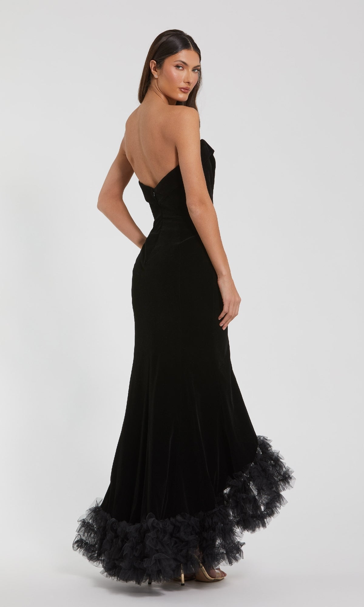 A woman models the Mac Duggal 68697 long formal dress, featuring a strapless velvet bodice, tulle ruffle hem detailing, and a thigh-high slit, as she poses looking over her shoulder against a plain background.